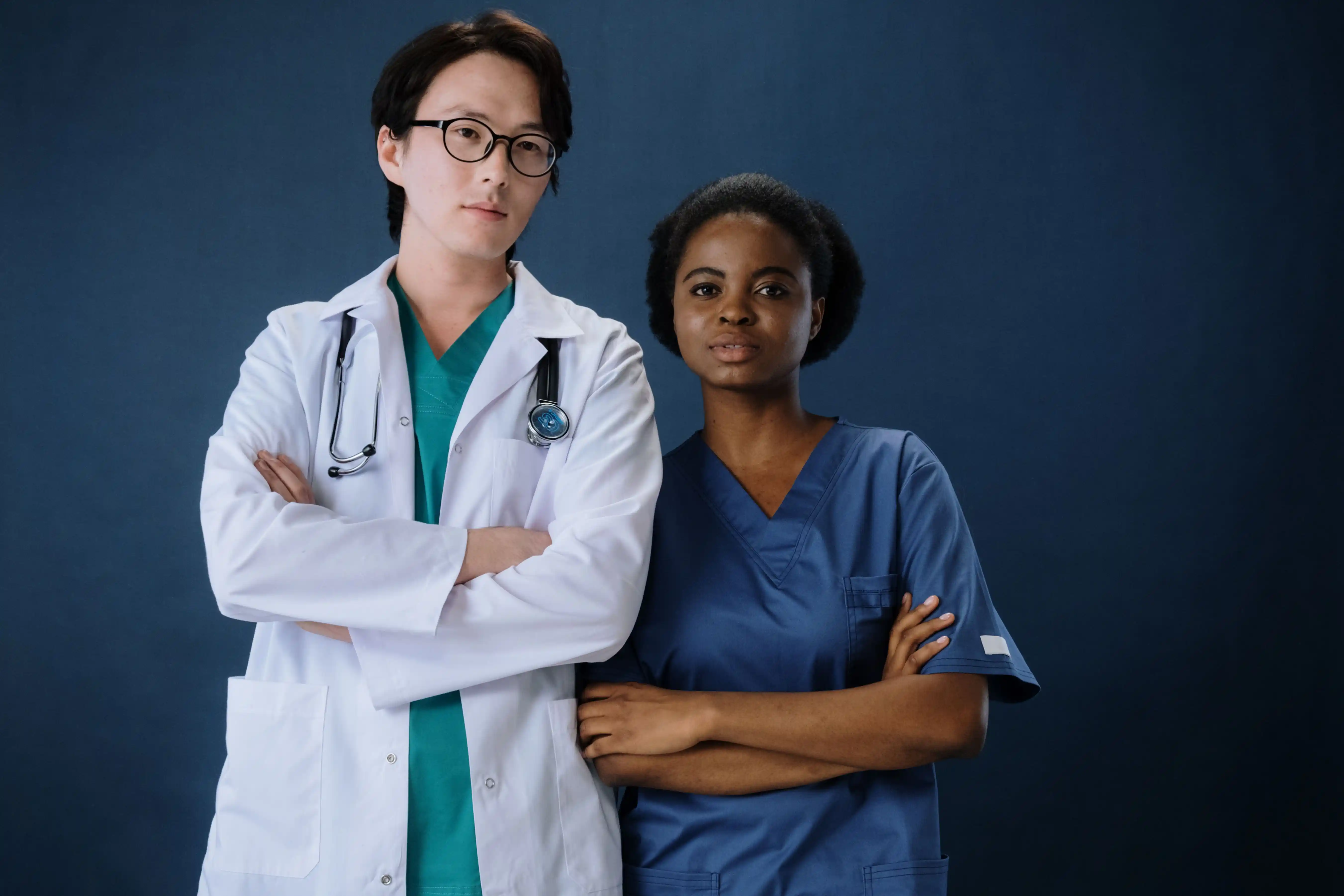 A doctor in a white coat and a nurse in blue scrubs standing side by side, arms folded